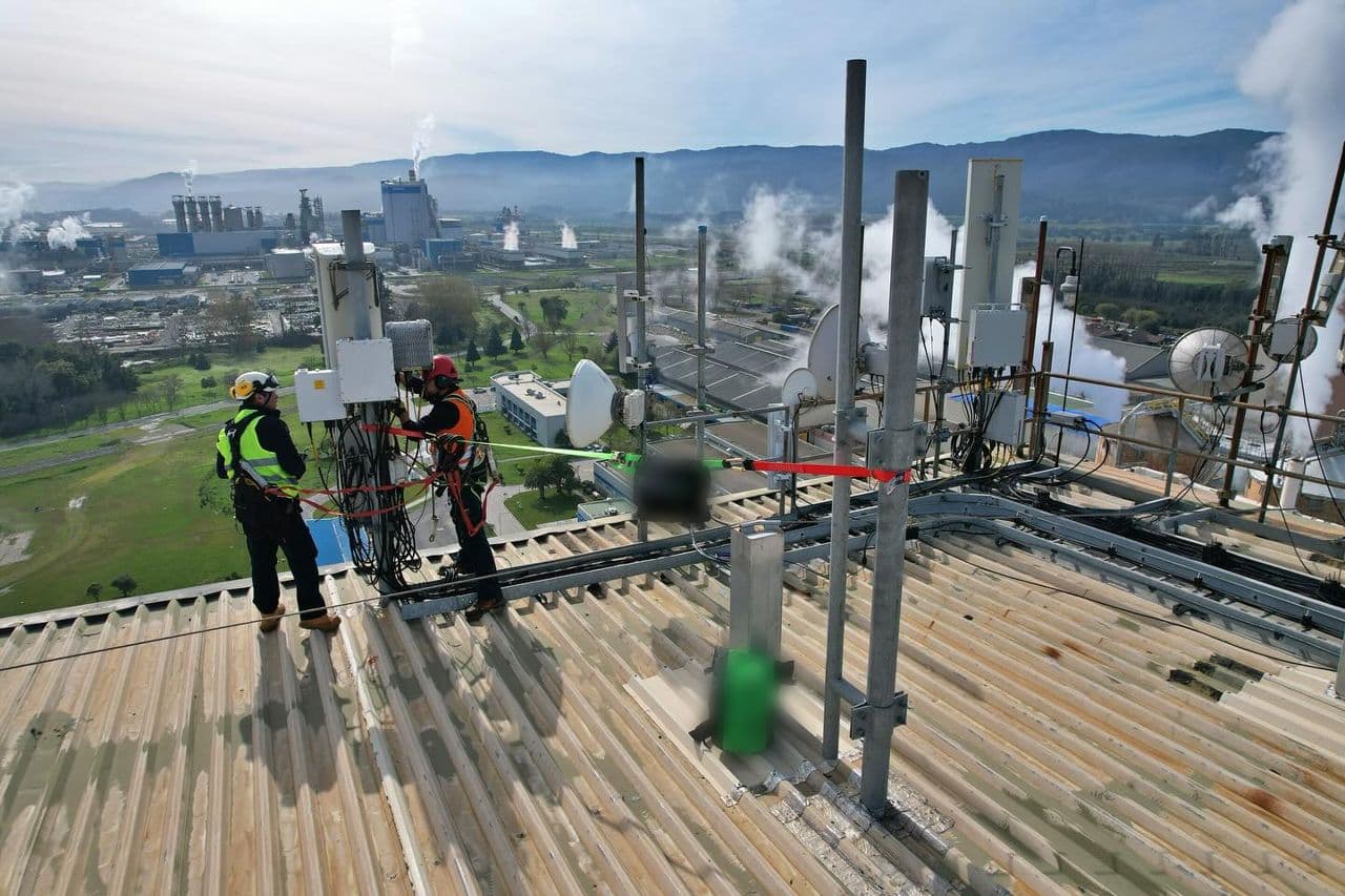 Dos técnicos con arnés trabajan junto a antenas de telecomunicaciones en la azotea de una planta industrial