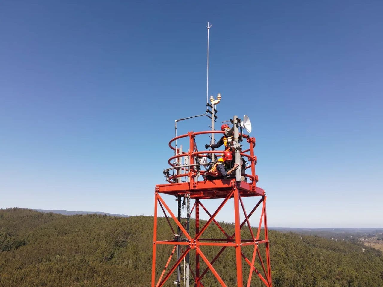 Dos técnicos en la plataforma superior de una torre de telecomunicaciones roja, sobre un bosque
